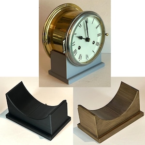 May include: A brass ship's clock with a white face and black Roman numeral hour markers. The clock is on a grey wooden base and is displayed on a black and a brown wooden stand.