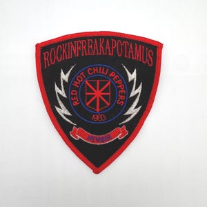 May include: A shield-shaped embroidered patch with a black background and red border. It features the red text ROCKINFREAKAPOTAMUS, a central red star emblem, the text RED HOT CHILI PEPPERS, the year 1983, white lightning bolts, and a red banner.