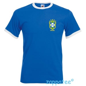 Pode incluir: Camiseta azul royal com acabamentos brancos na gola e mangas. A camisa apresenta o emblema da bandeira brasileira no peito esquerdo. O emblema inclui um escudo, estrelas e o texto "BRASIL".