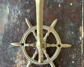 Puede incluir: Un gancho de metal dorado en forma de rueda de barco con un diseño decorativo. El gancho está fijado a una pared de madera.