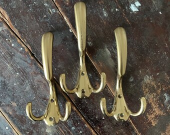 May include: Three brass coat hooks with a triple hook design. The hooks have a curved, elongated top and a decorative base with three screw holes. The hooks are displayed on a weathered, dark wood surface.