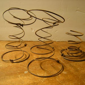 May include: Three antique metal coil springs of varying sizes, arranged on a wooden surface. The springs are a rusty brown color, with a spiral design. The largest springs have a circular top and bottom, while the smallest is a compact coil.