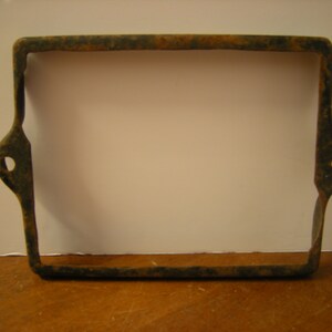 May include: Rectangular, rusty metal frame with rounded corners and two side tabs with circular holes. The frame has a dark, aged appearance, suggesting it is an antique or vintage item. The metal shows signs of oxidation.