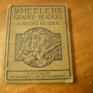 May include: A vintage book titled "Wheeler's Graded Readers - A Second Reader" with a black and white illustration of a child reading in a tree on the cover. The book is bound in brown fabric with black lettering.