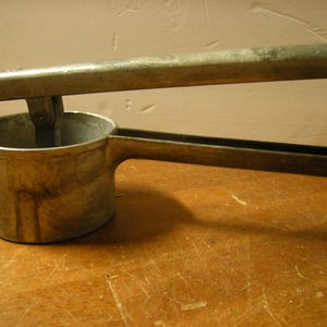 May include: A vintage, silver-colored potato ricer with a cup-shaped base, a long handle, and a lever. The ricer is made of metal and sits on a wooden surface. The handle is curved for easy gripping.