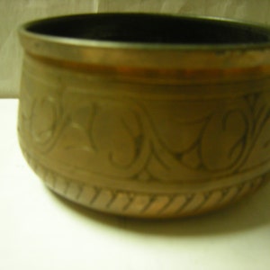 May include: A small, round, copper bowl with an intricate floral design etched into the surface. The bowl has a dark brown interior and a slightly raised rim.