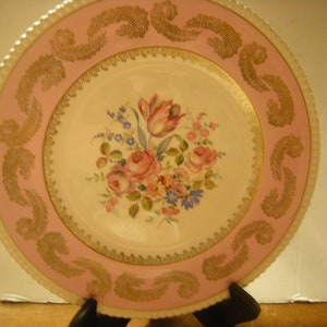 May include: A vintage porcelain dinner plate with a pink rim and gold trim. The center features a floral design with pink, yellow, and blue flowers.