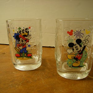 May include: Two clear glass tumblers featuring Mickey Mouse. One tumbler shows Mickey with a film clapper, the other with fireworks. Both glasses have the year 2000 printed on the base. The glasses have colorful designs and are suitable for cold beverages.