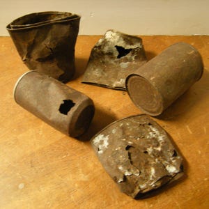 May include: An assortment of five rusted metal cans, some with holes and torn edges, arranged on a wooden surface. The cans vary in shape and size, displaying a weathered, brown color.