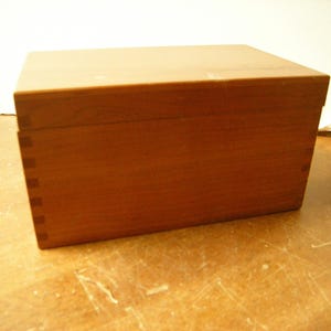 May include: A rectangular wooden box with a warm, reddish-brown hue. The box is constructed with visible dovetail joints at the corners, indicating a traditional woodworking technique. The lid sits flush with the box's sides.