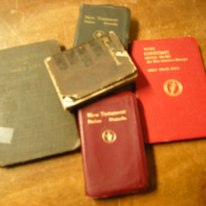 May include: A collection of small, vintage books in various colors, including green, brown, and red. Some books have gold lettering and religious symbols. The books are stacked and arranged on a wooden surface.