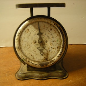 May include: An antique metal scale with a round face and a small platform. The face of the scale is white with black markings and a needle. The scale has a weathered, aged appearance.