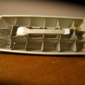 May include: A vintage metal ice cube tray with a silver finish. The tray has a rectangular shape with dividers and a lever marked "Magic Touch". The tray is sitting on a wooden surface.
