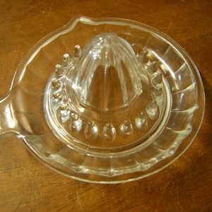 May include: A clear glass citrus juicer with a ridged base and a central cone with ridges. The juicer has a spout on the side for pouring juice.