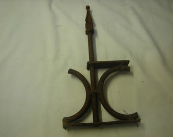 May include: A rusty metal fence post finial with a pointed top and two curved arms. The finial is made of iron and has a weathered, aged look.