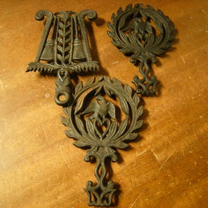 May include: Three antique cast iron trivets. The trivets are dark brown and feature ornate designs. One has a lyre shape, another has a wreath with an eagle, and the third has a similar wreath design. They are displayed on a wooden surface.