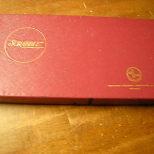 May include: A red Scrabble game box with gold lettering and a gold circular logo. The box is rectangular and sits on a wooden surface. The game box has a textured surface and is closed.