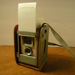 May include: Vintage Anscoflex camera in a green leather case with a matching strap. The camera has a silver top with the name "ANSCOFLEX" printed on it. The camera is a light green color with a silver lens.