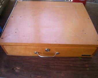 May include: A light brown wooden box with a drawer. The box has a metal handle and a keyhole on the drawer. The top of the box is a lighter shade of brown than the drawer. The box is sitting on a dark brown wooden surface.