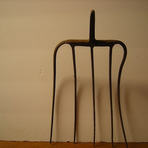 May include: A vintage, weathered metal pitchfork with four tines and a handle. The pitchfork is a dark, rusty brown color, suggesting age and use. The tines are long and slightly curved, and the handle has a vertical extension.