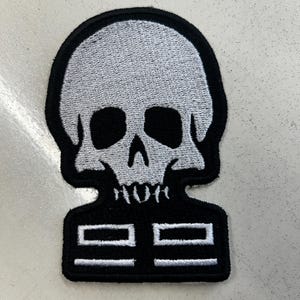 May include: An embroidered patch featuring a stylized light gray skull with black eye sockets and a nose cavity, positioned above a black rectangular base with four white horizontal rectangular outlines arranged in two rows of two.