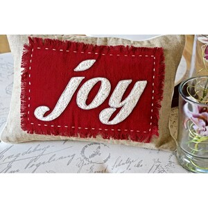 Könnte beinhalten: Ein rechteckiges Kissen aus beigem Leinen. Ein rotes Stofffleckchen mit dem Wort "JOY" in weißen, strukturierten Buchstaben ist auf das Kissen genäht. Der rote Patch hat einen ausgefransten Rand und weiße Nähte.