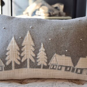 May include: Gray rectangular decorative pillow with a winter scene. The pillow features a landscape with trees and houses in a light beige color, with small, sparkling embellishments. The background is a darker gray.