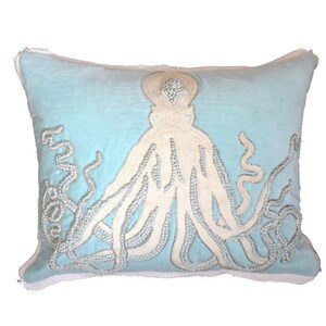 Puede incluir: Una almohada de color azul claro con un diseño de pulpo blanco con cuentas. El pulpo tiene ocho tentáculos y está centrado en la almohada.