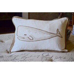 Puede incluir: Almohada blanca con un diseño de ballena bordado en marrón. La almohada tiene un ribete de arpillera marrón.