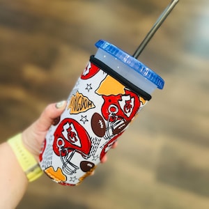 Puede incluir: Un vaso blanco con tapa azul y pajita negra. El vaso presenta un patrón repetido de cascos de fútbol americano rojos, balones de fútbol y la palabra "KINGDOM" en amarillo. Una banda negra rodea el centro.