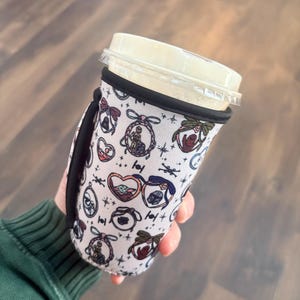 May include: A white coffee cup sleeve with a black handle and trim. The sleeve features a repeating pattern of cartoon characters in heart-shaped frames, bows, and stars. The cup has a clear plastic lid and is held by a person wearing a green long-sleeve shirt.