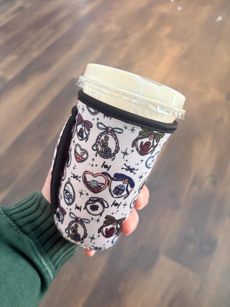 May include: A white coffee cup sleeve with a black handle and trim. The sleeve features a repeating pattern of cartoon characters in heart-shaped frames, bows, and stars. The cup has a clear plastic lid and is held by a person wearing a green long-sleeve shirt.