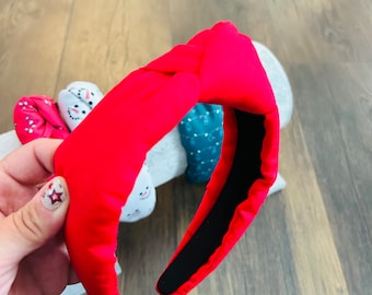 May include: A red fabric headband with a knotted design. The headband is made of a soft, plush material and has a black elastic band on the inside.