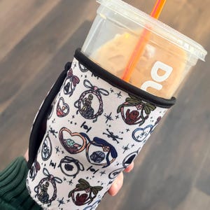 May include: A white and black drink sleeve with a Star Wars-themed pattern, holding a clear plastic cup with a straw. The sleeve features various character illustrations and a black handle.
