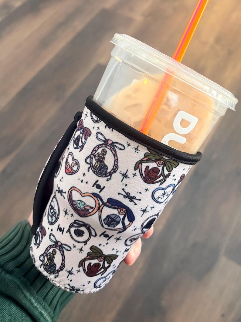 May include: A white and black drink sleeve with a Star Wars-themed pattern, holding a clear plastic cup with a straw. The sleeve features various character illustrations and a black handle.