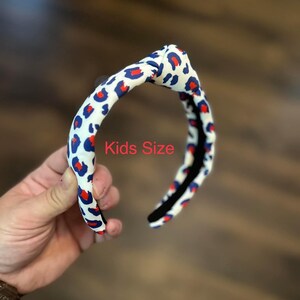 May include: A white headband with a red and blue leopard print pattern. The headband is knotted on top and says "Kids Size" in red text.