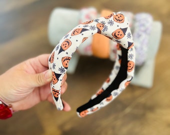 May include: A white headband with a black and orange print of pumpkins and witch hats. The headband is made of a soft, stretchy material and is perfect for adding a touch of Halloween fun to any outfit.