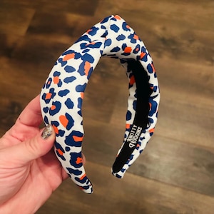 May include: A white headband with a blue and orange leopard print. The headband is knotted on top and has the text "Knotty Noggins" printed on the inside.