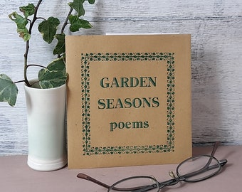 May include: A brown paper book with a green border and the text "GARDEN SEASONS poems" on the cover. The book is sitting on a light brown surface with a pair of glasses and a vase with green leaves.