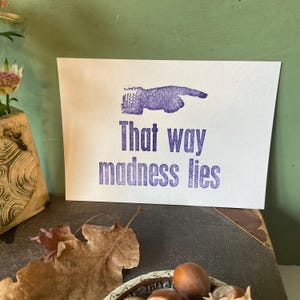 May include: A white card with purple text and a hand pointing to the right. The text reads "That way madness lies." The card is on a dark surface with leaves and a bowl of nuts.