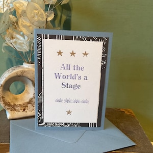 May include: A greeting card with the text "All the World's a Stage" in purple script, adorned with star and floral designs. The card is set against a light blue background and comes with a matching envelope. A decorative vase with dried flowers is in the background.