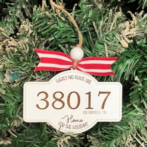 May include: White Christmas ornament with the text "There's no place like 38017 Collierville, TN Home for the Holidays." The ornament has a red and white striped ribbon and a wooden bead. It hangs from a twine string.
