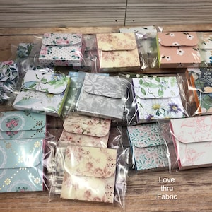 May include: A collection of small, square, fabric-covered boxes with a flap closure. The boxes are arranged in rows and are packaged in clear plastic bags. The boxes feature various floral and patterned designs, including a floral pattern with a white background, a gray pattern with a floral design, and a pink pattern with a floral design. The text "Love thru Fabric" is visible at the bottom of the image.
