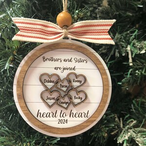 May include: A wooden Christmas ornament with a white background and a brown wood frame. The ornament features the text "Brothers and Sisters are joined heart to heart 2024" and five cutout hearts with names inscribed inside: Debbie, Ronny, Tricia, Jason, and Becky.