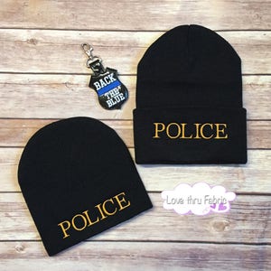 May include: Two black knit beanies with the word "POLICE" embroidered in gold on the front. A keychain with a blue and white "Back the Blue" patch is also pictured.
