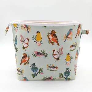 May include: A light green zippered pouch with a colorful bird pattern. The birds are depicted in various colors and poses, with floral accents. The pouch has a white zipper and a white interior lining. The pouch has a rectangular shape.