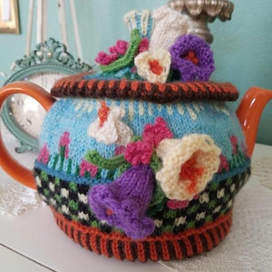 May include: A ceramic teapot with an orange handle and spout, adorned with a knitted cozy. The cozy is light blue with colorful knitted flowers in pink, purple, and cream. The cozy has a brown and orange trim.