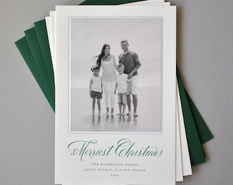Op de afbeelding: Een stapel kerstkaarten met een zwart-wit familiefoto. De kaarten hebben een witte rand en de woorden "Merriest Christmas" in groen schrift. Groene enveloppen zijn zichtbaar.