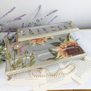 May include: Two-tiered wooden tray with a gray painted finish and floral design. The top tier has a gold trim and a lavender floral design. The bottom tier has a sunflower and rose floral design. The tray has gold metal accents and decorative feet.