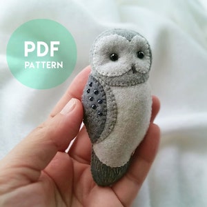 Può includere: Un gufo di feltro grigio con occhi neri e pancia bianca. Il gufo è alto circa 12,5 cm ed è tenuto in una mano. Il gufo è un modello PDF.